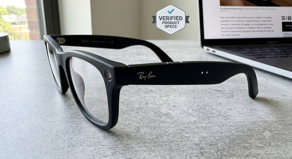 Black Ray-Ban Meta smart glasses on a grey desk, showing the side logo and camera, with a blurred laptop and a specs badge.
