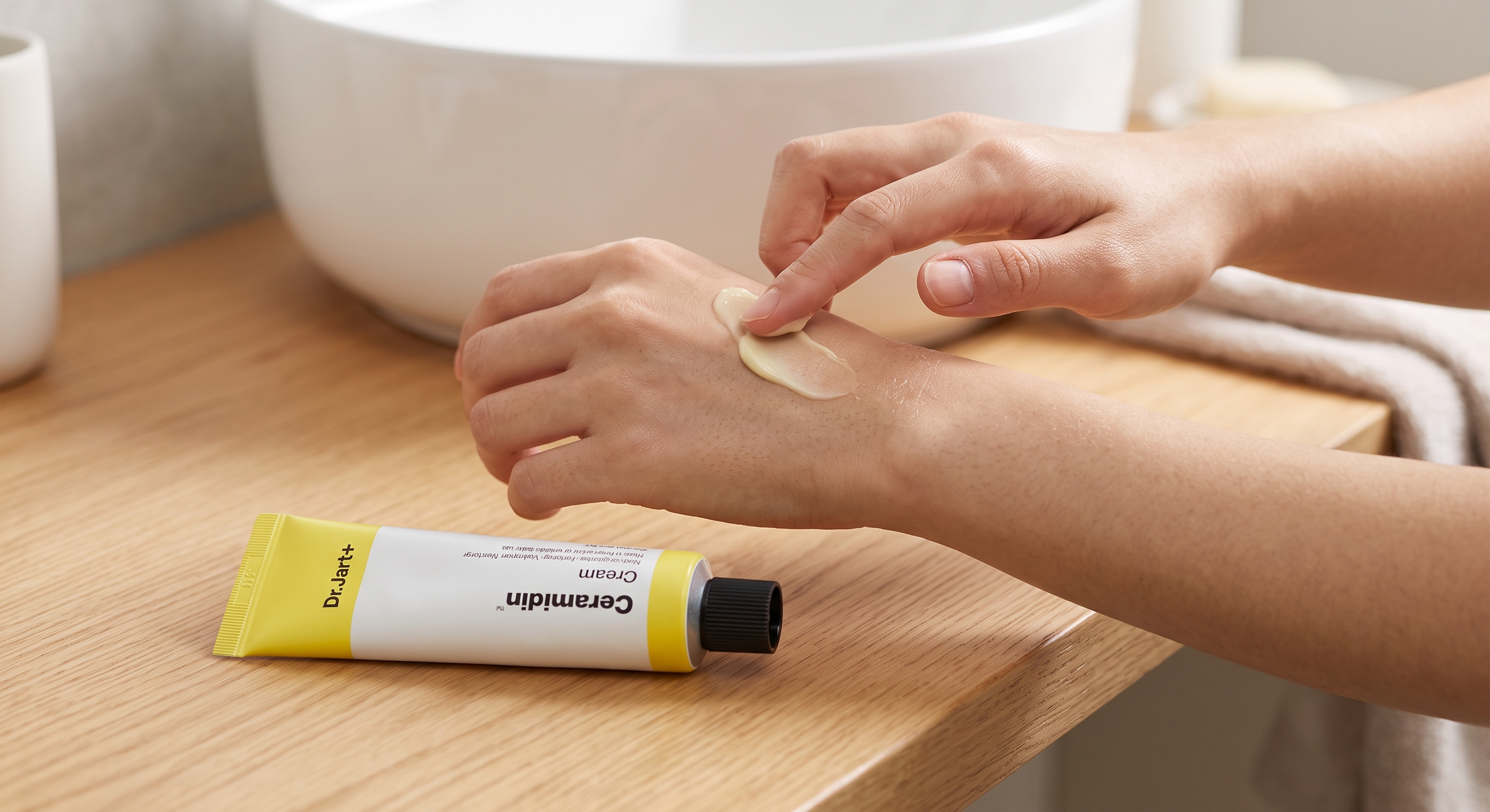 Hands rub a thick, creamy dollop of Dr.Jart+ Ceramidin Cream into the skin next to the yellow tube on a wooden counter.