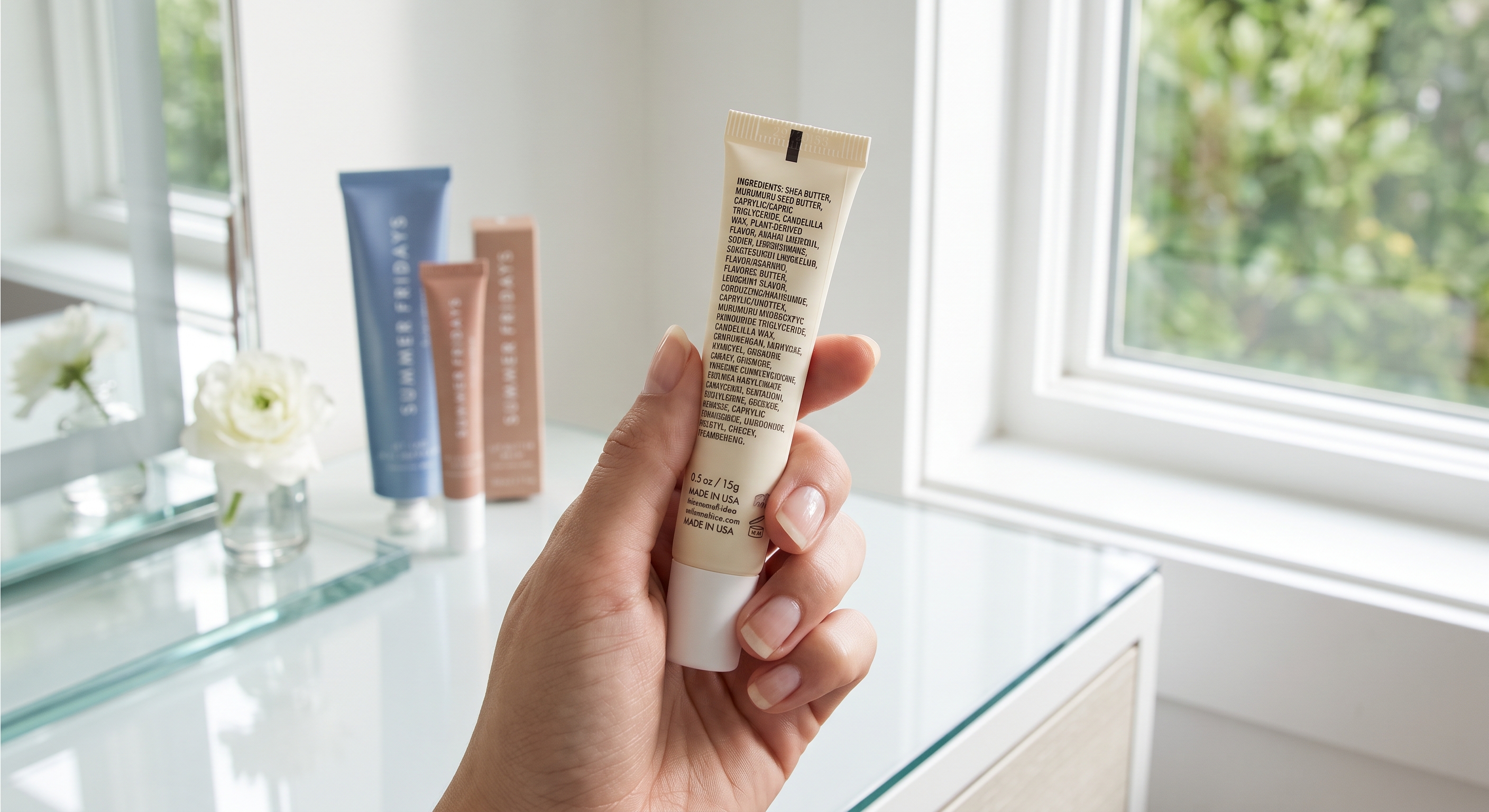 Hand holds a small tube, reading ingredients, with beauty products and white flower on a glass table by a window.