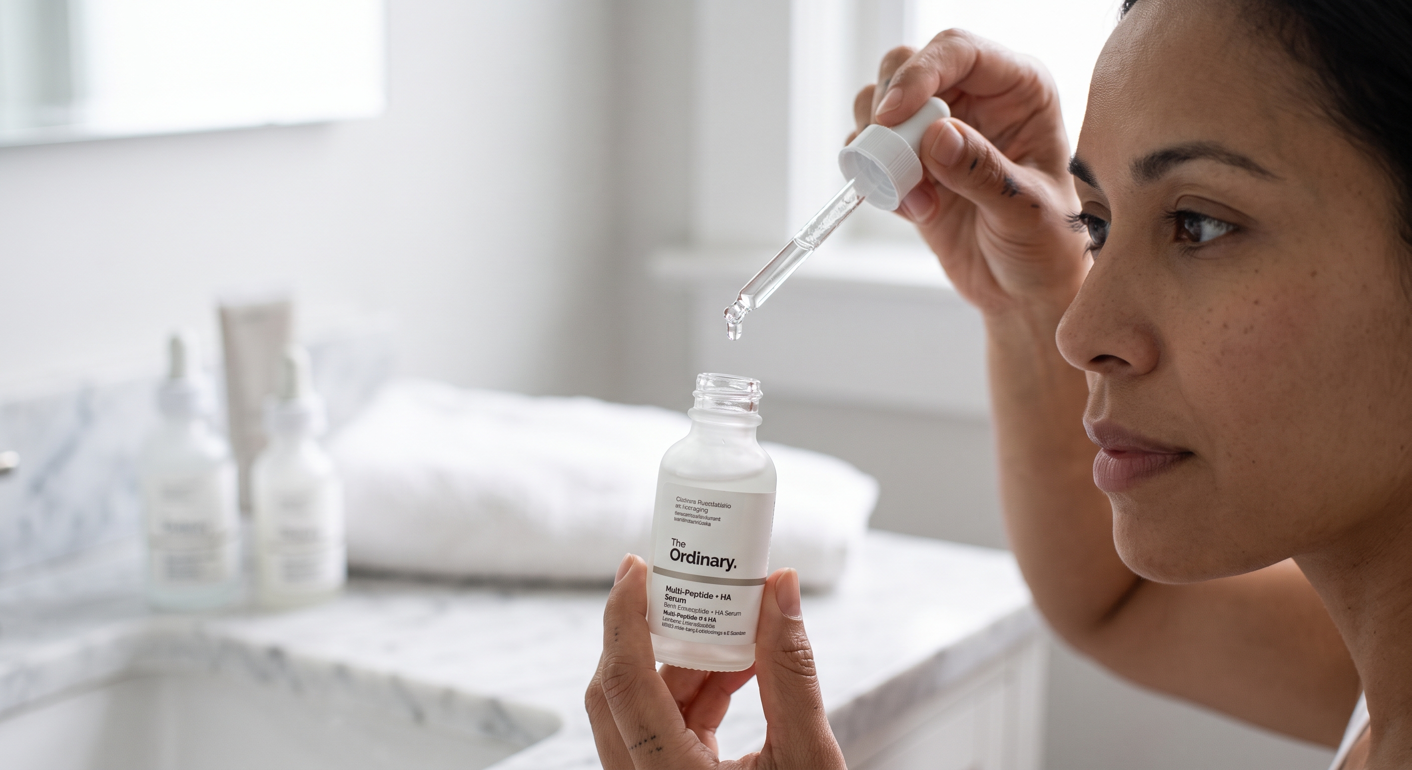 A woman holds a bottle of The Ordinary Multi-Peptide + HA Serum and a glass dropper in a brightly lit bathroom.
