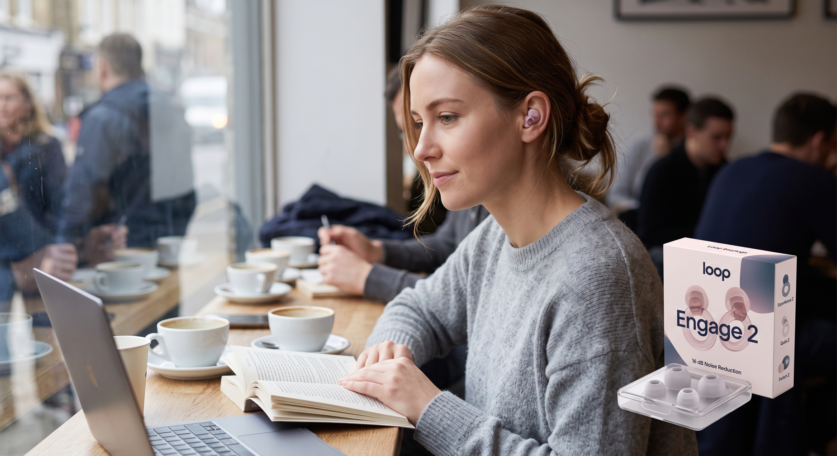 A woman wears Loop Engage 2 earplugs while reading in a busy cafe, a branded box of earplugs sits on her table.