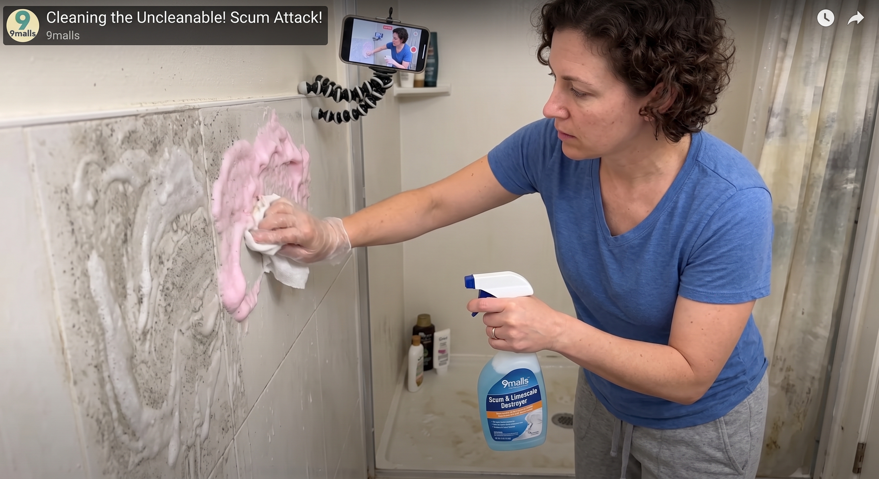 A woman scrubs pink foam from bathroom tiles with a gloved hand and spray.