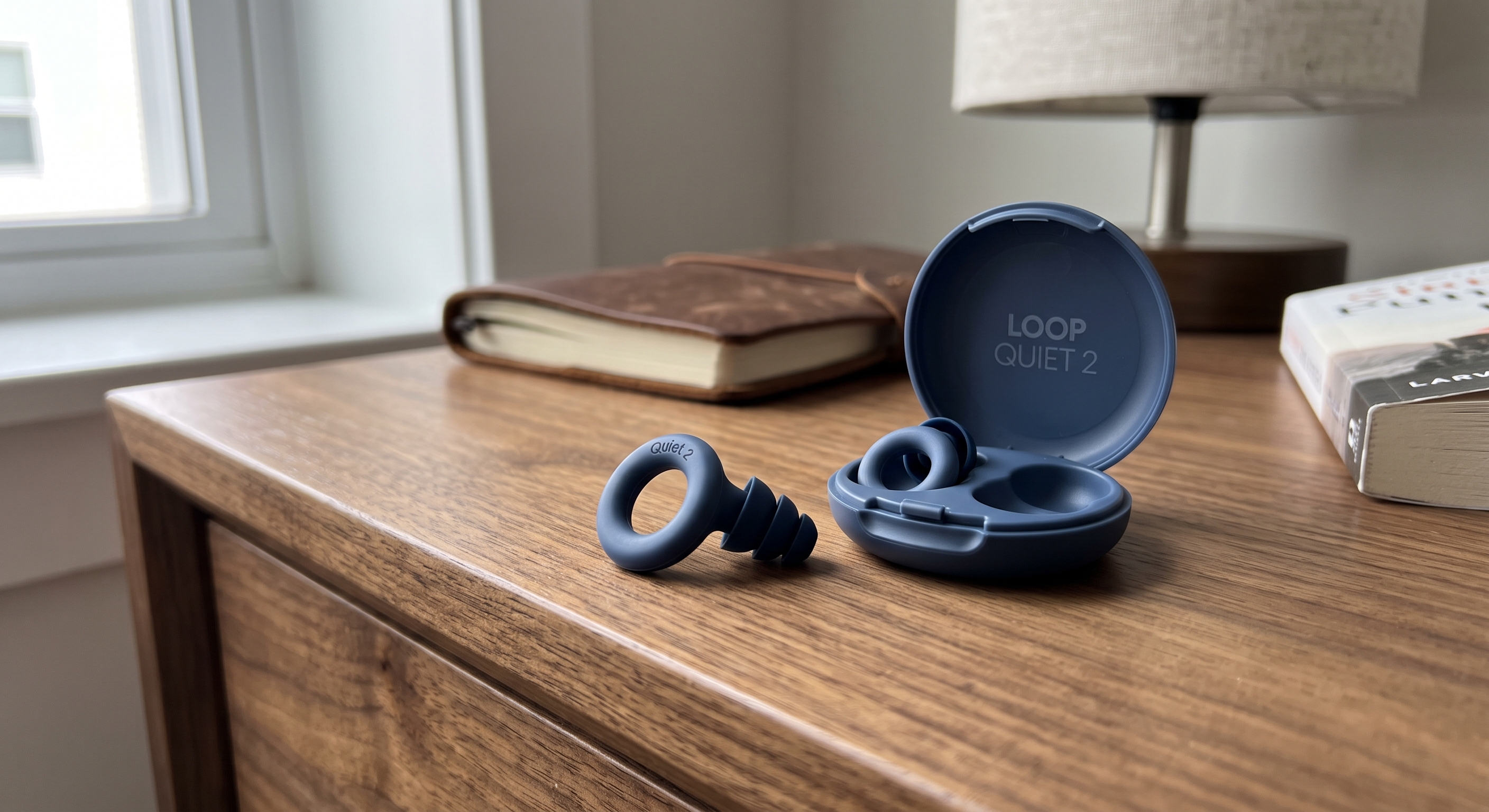 A dark blue Loop Quiet 2 earplug rests on a wooden nightstand next to its matching open carrying case with the same product name.