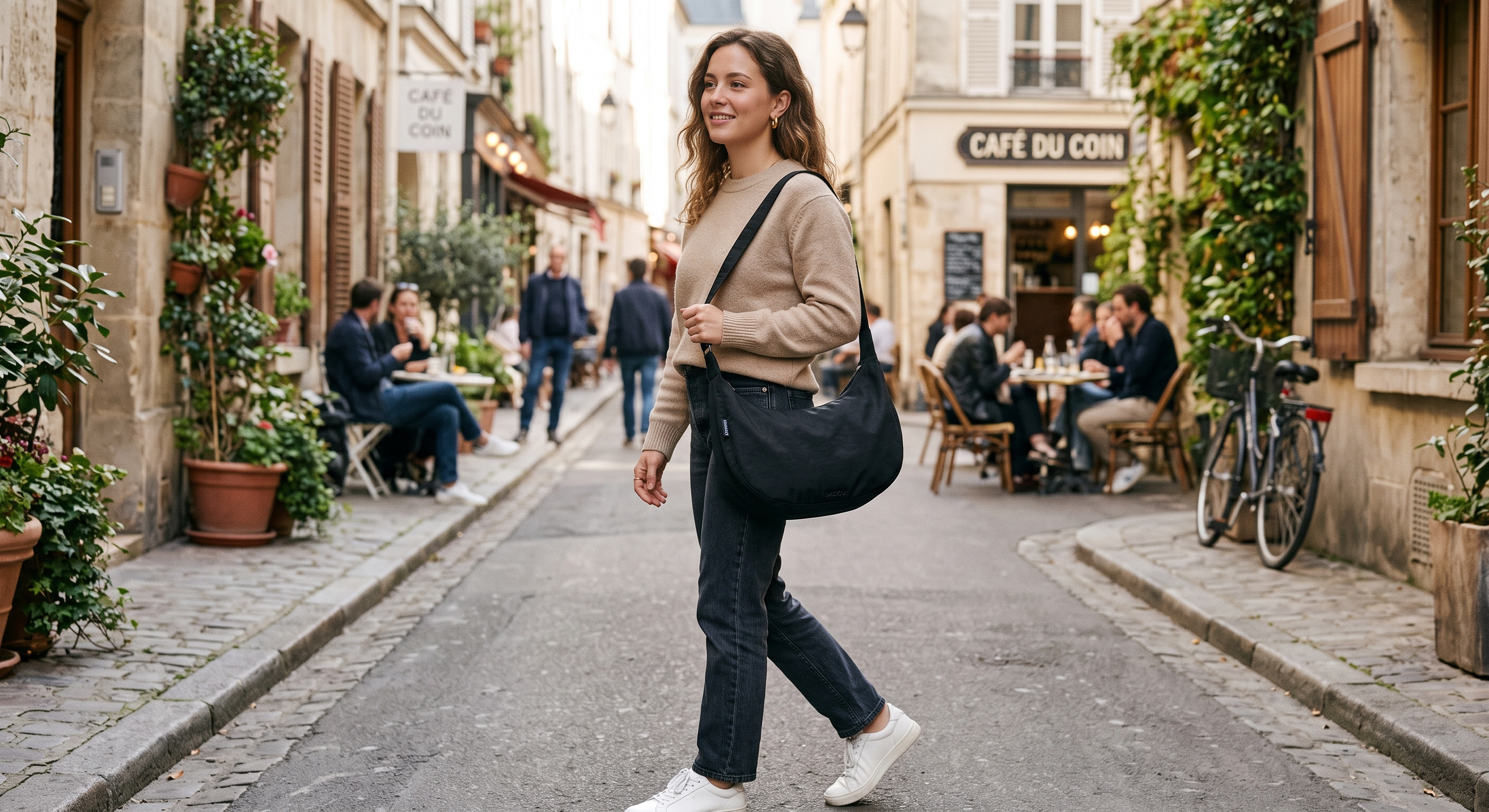A woman in a tan sweater walks down a European street carrying a black crossbody bag, with cafes in the background.